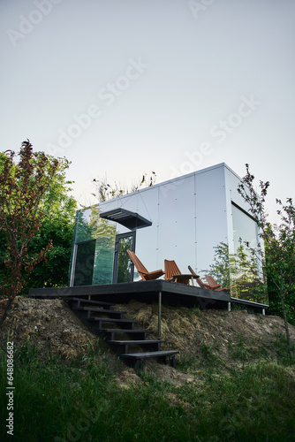 modern eco-friendly glass house with wooden stairs and chair on porch, tranquil countryside setting © LIGHTFIELD STUDIOS