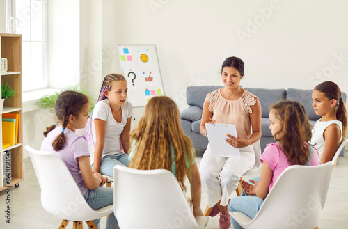 Young smiling woman psychologist conducts a mental health lesson for a group of school children girls sitting in a circle and having team work training during therapy session in classroom.