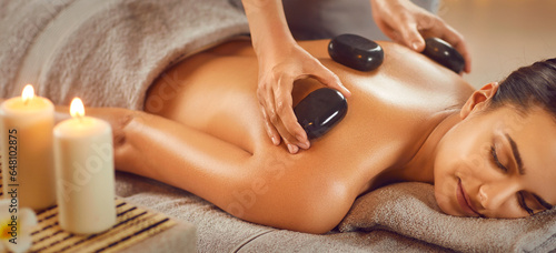Relaxed young woman enjoying body treatment at wellness center. Beautiful serene girl lying on couch getting hot stone massage in spa salon. Beauty treatment therapy, body care concept