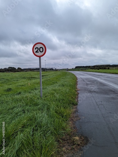 View of 20mph warning sign alongside low trafficked road, surrounded by grass and cloudy sky. photo