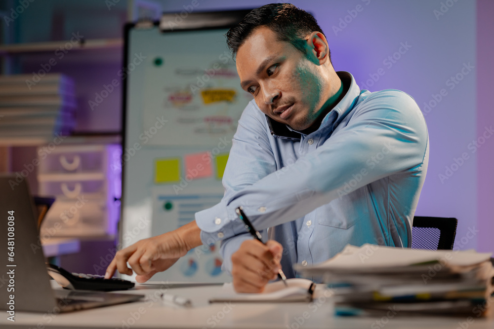 Asian man talking on the phone in the office of a startup company, company employees working overtime, late night work, overtime of a busy and hard working company employee. Overtime concept.