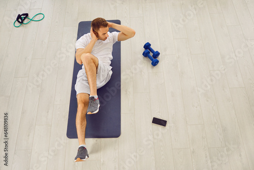 Overhead view from above of man doing criss cross crunches or sit ups exercise on rubber sports mat on white wooden gym floor with dumbbells, resistance band and mobile phone. Fitness workout concept