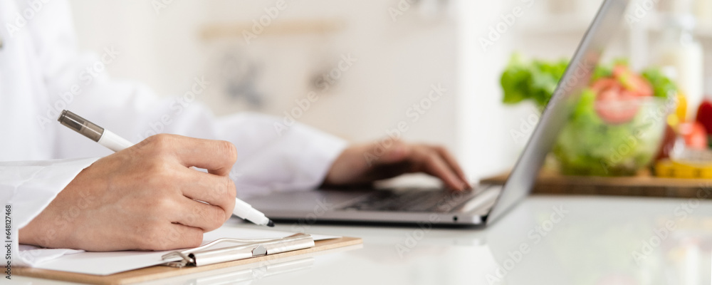 Close-up of dietitian hand writing with laptop giving online weight loss and healthy diet consultation with fresh vegetable fruit measuring tape and glass of clean water on desk against