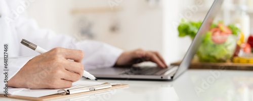 Close-up of dietitian hand writing with laptop giving online weight loss and healthy diet consultation with fresh vegetable fruit measuring tape and glass of clean water on desk against