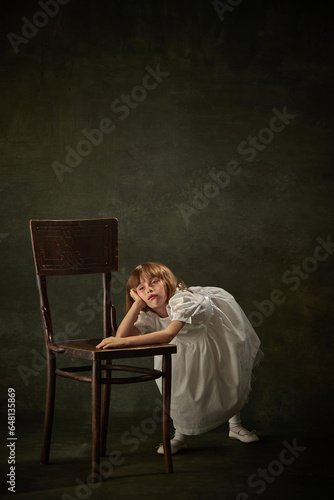Portrait of boring little girl, child in white dress leaned on chair against vintage background. Fairy princess or queen. The heroine of story tale.