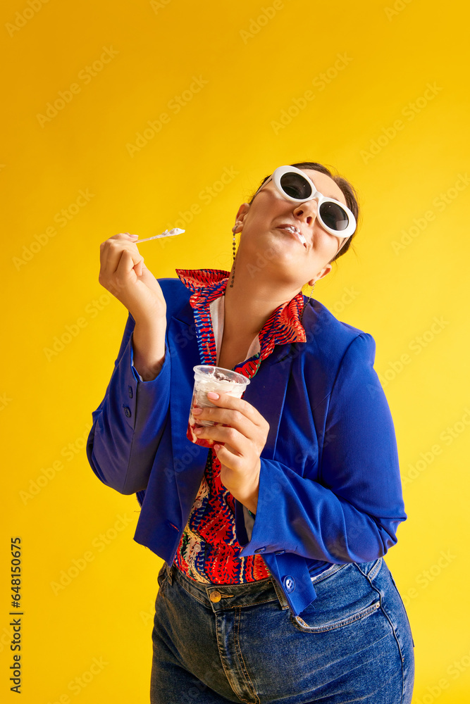 Portrait of stylish, elegant young woman in beautiful costume and sunglasses, eating ice cream against bright yellow studio background. Concept of beauty, human emotions, lifestyle, fashion, ad