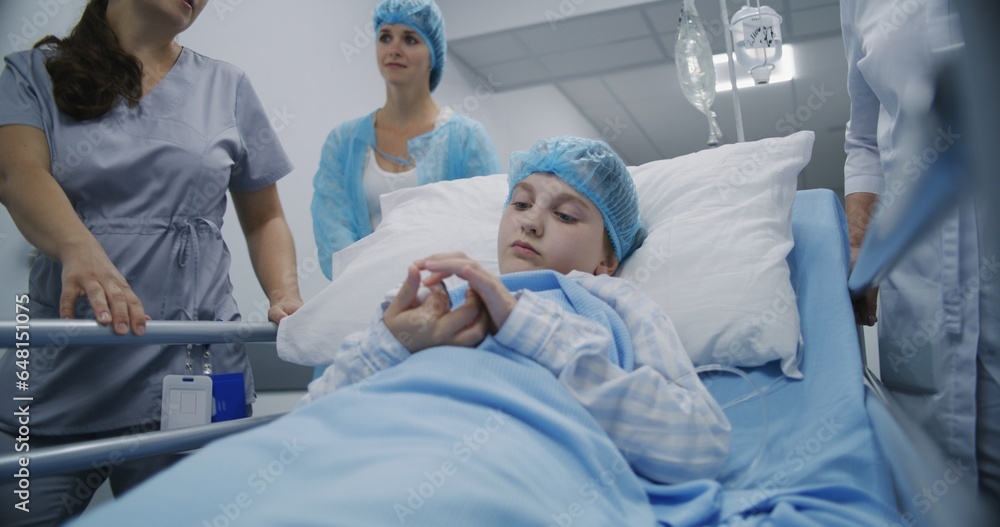 Obraz premium Medics transport patient to hospital room after surgery down medical center hallway. Young girl lies on gurney, looks at pulse oximeter on her finger and talks with mother. Medical staff at work.