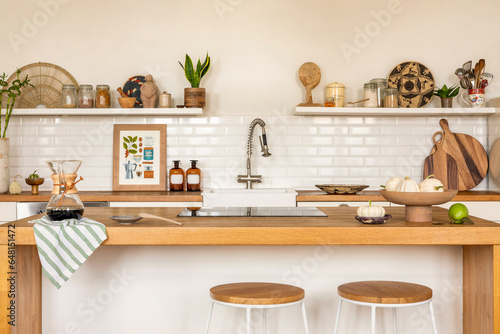 Creative composition of kitchen interior with mock up poster frame, wooden kitchen island, simple barstool, vase with green flowers, silver tap and personal accessories. Home decor. Template.