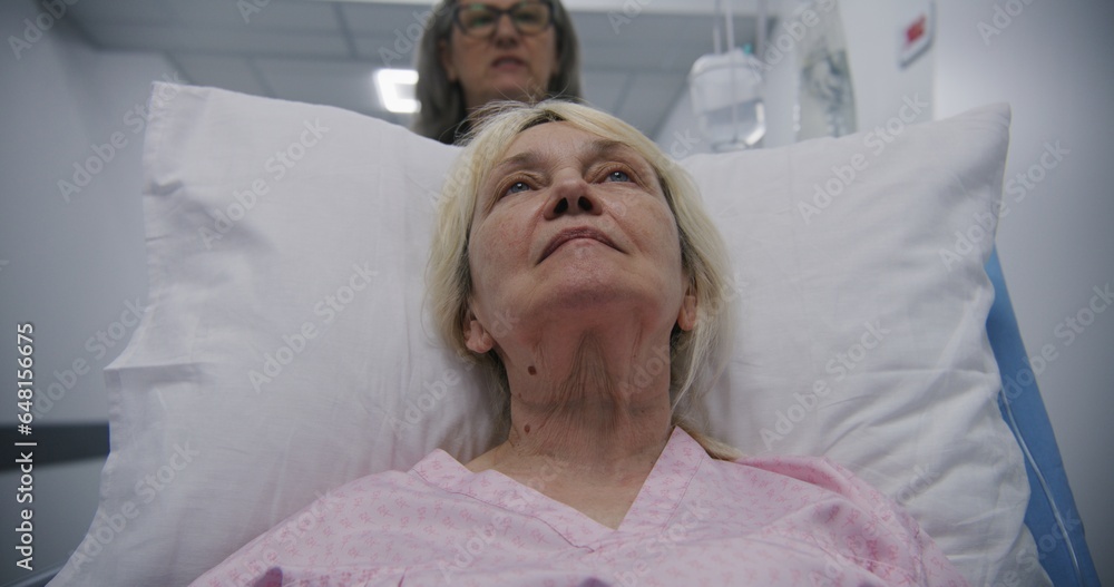 Fototapeta premium Senior woman lies on gurney, talks with mature female doctor. Physician transports patient to hospital ward down modern medical center hallway after surgery. Medical personnel at work in hospital.