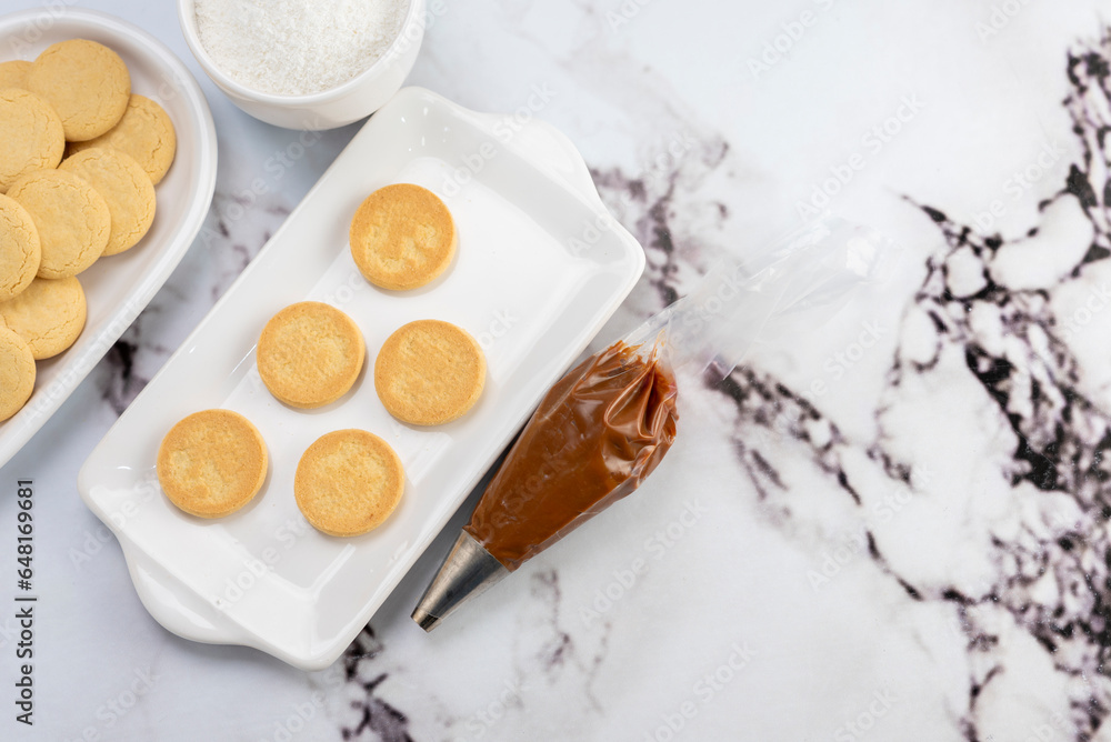 ingredients and mise en place for the preparation of cornstarch alfajores, filling the dulce de leche filling on top and grated coconut on the outside on white plates. ingredients and mise en place for the preparation of cornstarch alfajores, filling the dulce de leche filling on top and grated coconut on the outside on white plates.