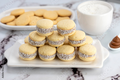 Homemade cornstarch alfajores filled with dulce de leche with coconut and cup of tea on plates and white background