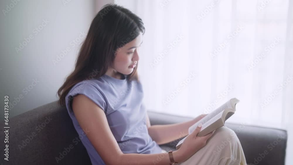 Attractive young Asian woman relaxed in casualwear reading book sitting on modern sofa in front of window relaxing in her living room relaxing concept. Attractive young Asian woman relaxed in casualwear reading book sitting on modern sofa in front of window relaxing in her living room relaxing concept.