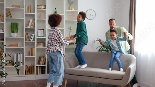 Multiracial family of mother, father and two preschool boys fooling around and spending active free time at home. Energetic kids holding parents' hands and jumping on couch. Leisure activity indoors. photo