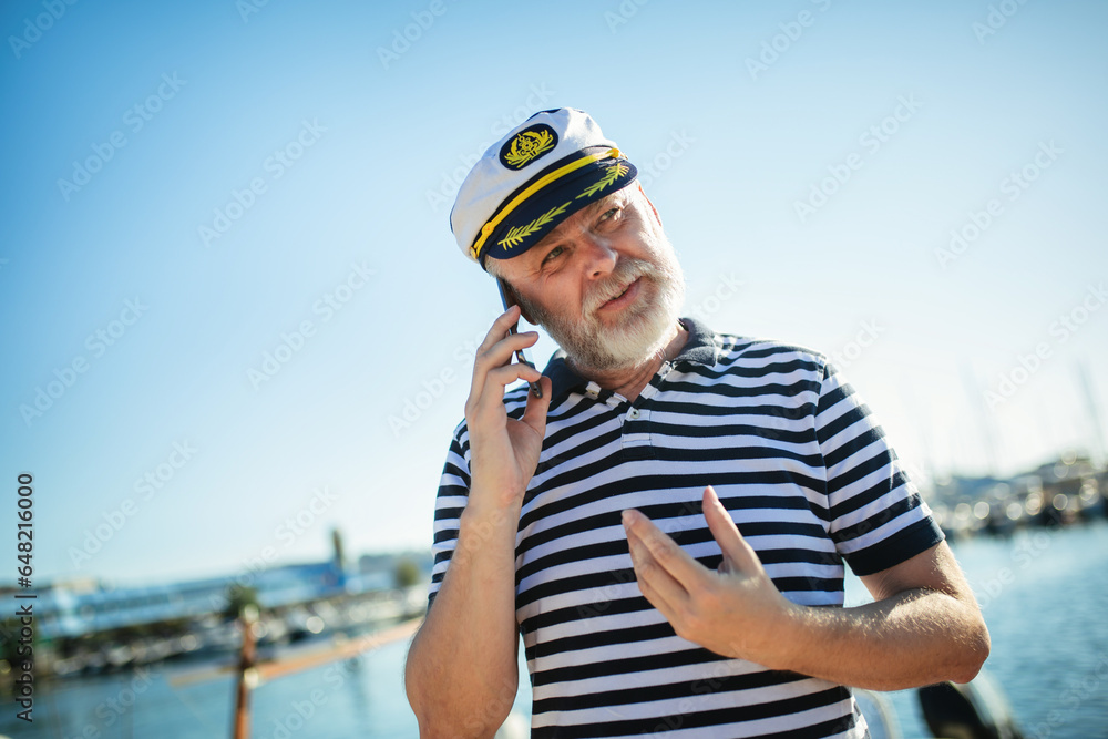 Mature man standing near the sea dressed in a sailor's shirt and hat using smart phone