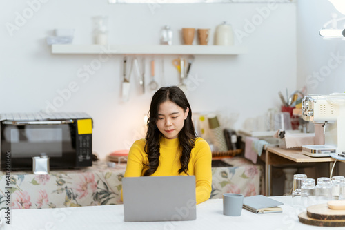 Beautiful Asian woman smiling happily relaxing using technology of laptop computer Take notes Drink a relaxing hot drink while sitting on the table in your room at home.