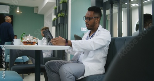African American doctor sits in clinic cafe, uses digital tablet. Professional medic consults patient via video call, has online appointment. Medical staff in hospital or medical center dining room.