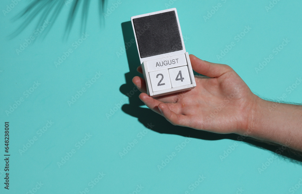Man's hand holding wooden block calendar with date august 24 on blue background with palm leaf shadow Man's hand holding wooden block calendar with date august 24 on blue background with palm leaf shadow