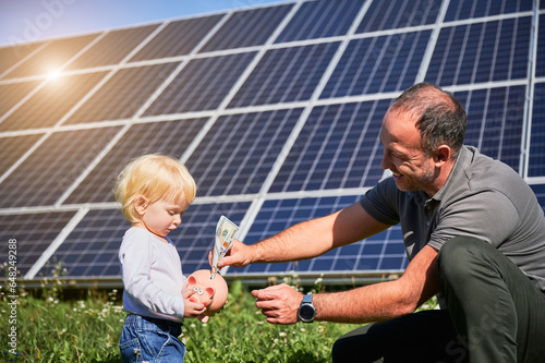 Father and his little child putting cash into piggy bank to save money on background of solar panels. Little kid interested in saving money for future. Concept of investment in renewable energy.