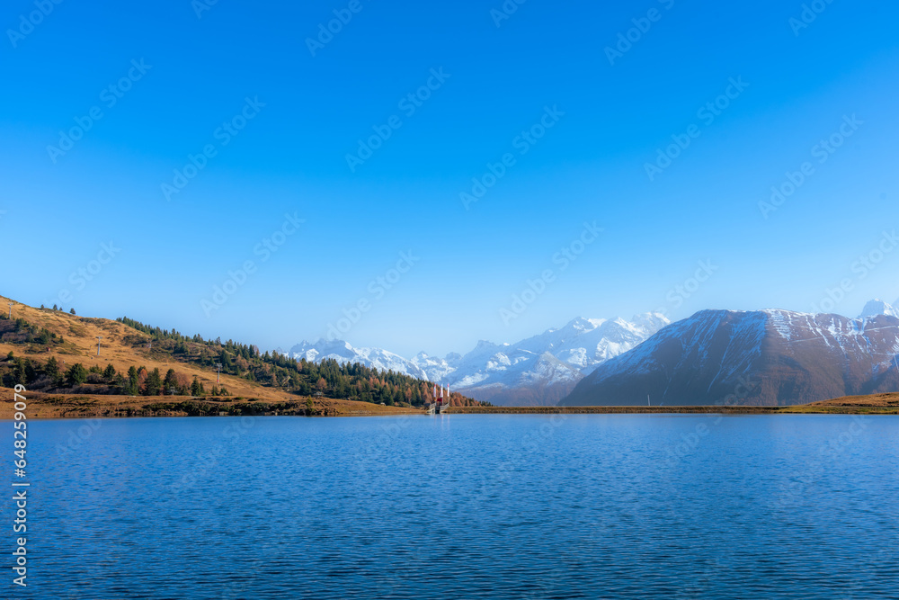 Scenic view of a lake, an autumn forest and mountains with snowy peaks.