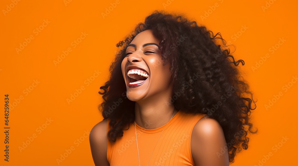 In front of an orange studio backdrop, a young black woman with long, curly hair and wearing a bra laughs while standing sideways. In front of an orange studio backdrop, a young black woman with long, curly hair and wearing a bra laughs while standing sideways.
