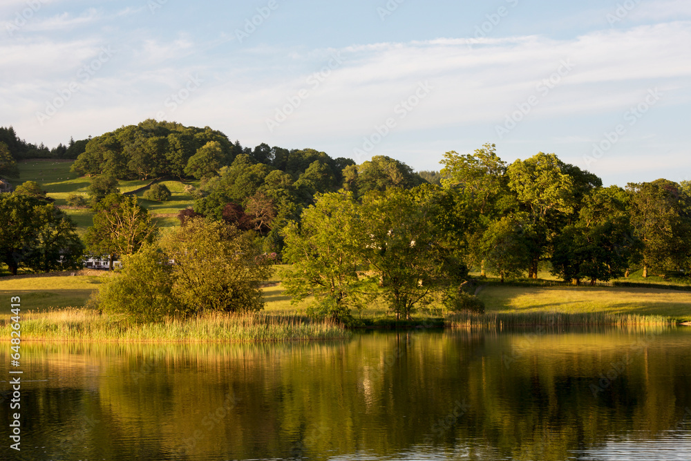 Tranquil Water Of A Lake Reflected The Green Trees And Grass Fields; Lake District, Cumbria, England Tranquil Water Of A Lake Reflected The Green Trees And Grass Fields; Lake District, Cumbria, England
