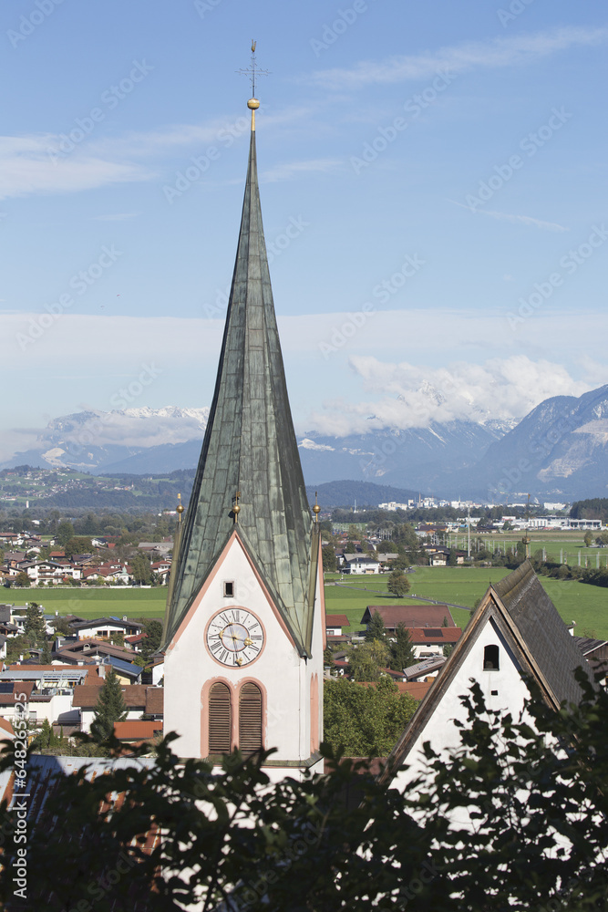 Church Steeple In A Village With Mountain Range, Blue Sky And Clouds In The Distance; Rattenburg, Austria Church Steeple In A Village With Mountain Range, Blue Sky And Clouds In The Distance; Rattenburg, Austria
