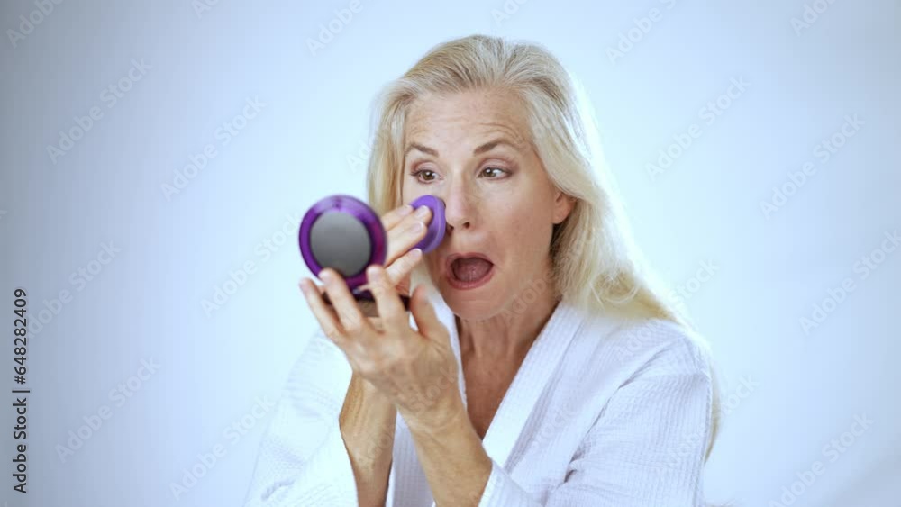 Portrait of 50s, 60s, mature middle aged woman applying natural makeup powder foundation with cosmetic sponge on clean face skin smoothing wrinkles, looking in mirror beauty routine white background. Portrait of 50s, 60s, mature middle aged woman applying natural makeup powder foundation with cosmetic sponge on clean face skin smoothing wrinkles, looking in mirror beauty routine white background.