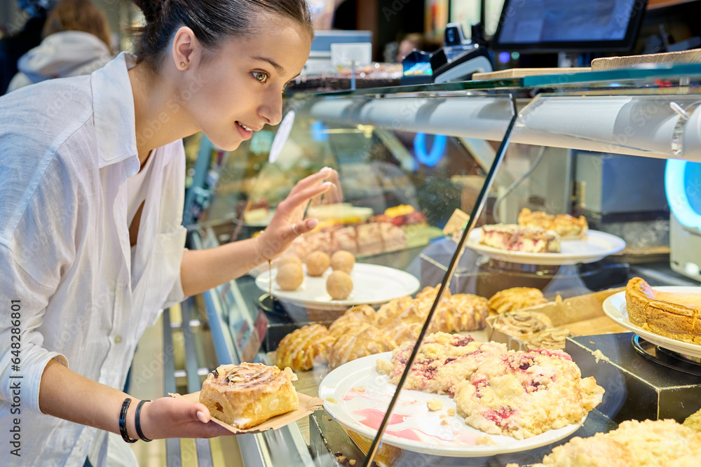 Young teenage female looking at sweet buns in glass display case in pastry shop