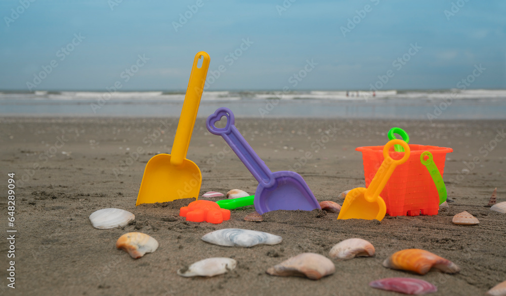 Beach toys, shovels and bucket on the sand surrounded by shells with the sea and blue sky in the background Beach toys, shovels and bucket on the sand surrounded by shells with the sea and blue sky in the background