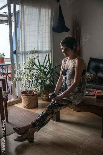 Mature woman sitting thoughtfully at the coffee table in her living room with her legs stretched out and a cup of tea in her hand. photo