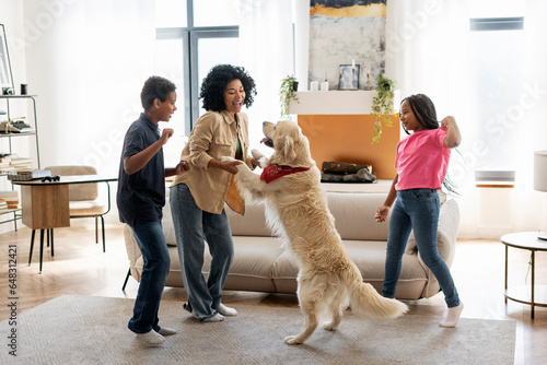Smiling African American family, mother, daughter and son playing dancing with golden retriever