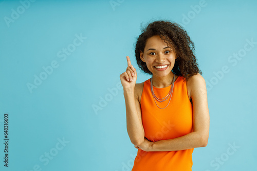 Woman in orange dress pointing finger up with smile on blue studio background. Advertising concept