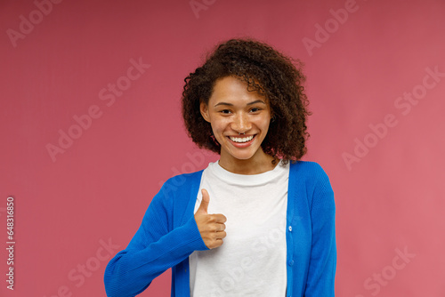 Cheerful woman in casual clothes showing thumb up with smile standing on pink studio background