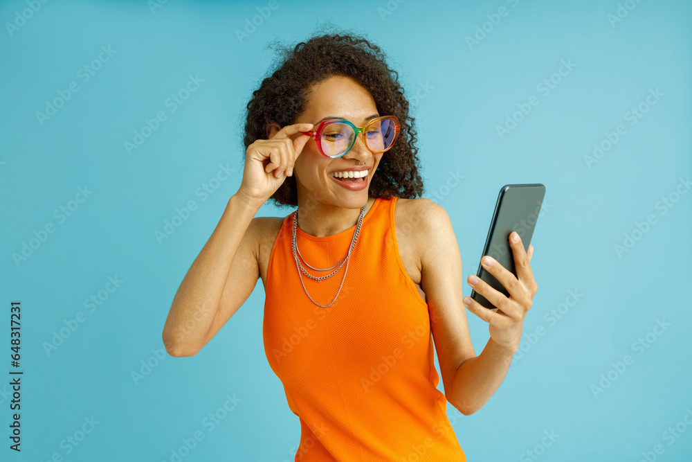 Smiling woman in eyeglasses looking on mobile phone while standing on blue studio background