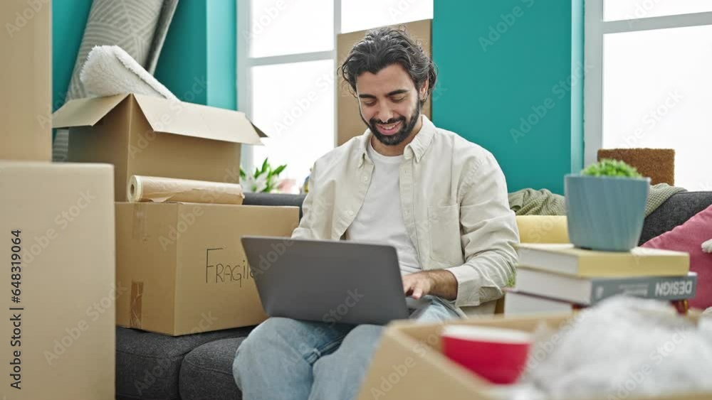 Young hispanic man using laptop sitting on sofa at new home Young hispanic man using laptop sitting on sofa at new home