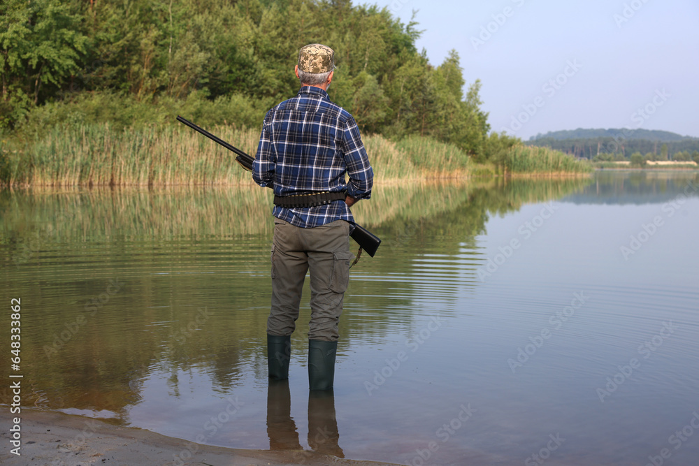 Man with hunting rifle near lake outdoors, back view Man with hunting rifle near lake outdoors, back view
