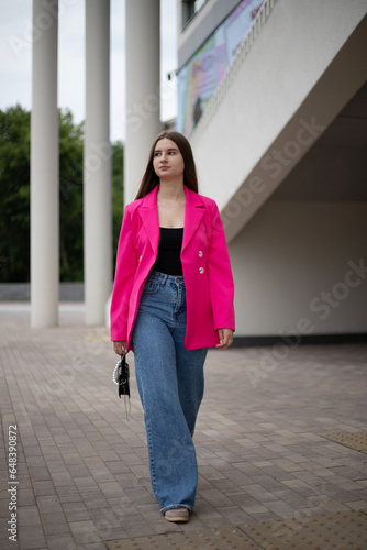 a young business girl in a bright fuchsia jacket and jeans walks on the street with a small handbag