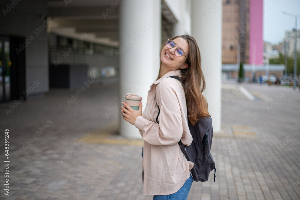 a young beautiful girl student in jeans and a shirt with a backpack and a mug of coffee on the street walks along the campus