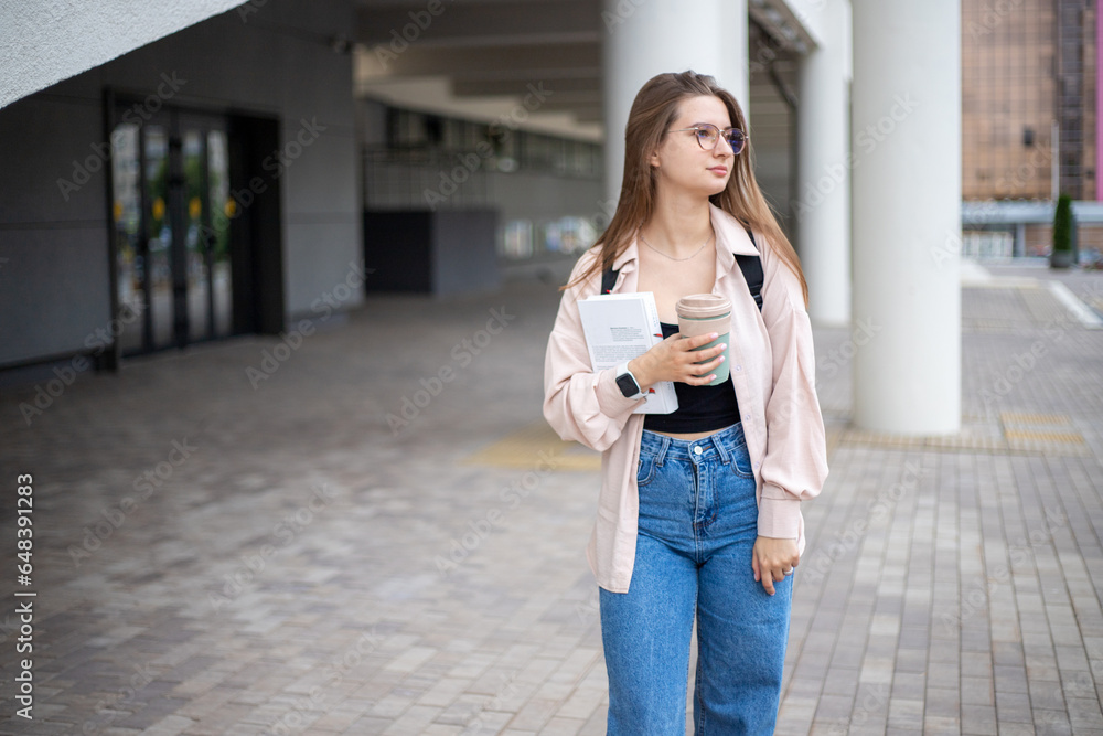 a young girl student in jeans and a shirt with a backpack and books and a mug of coffee on the street walks along the campus