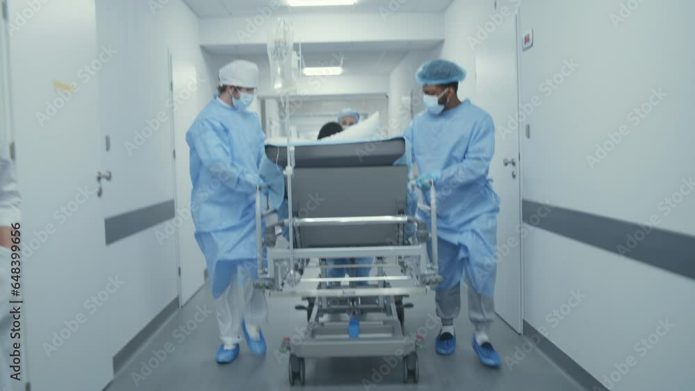 Doctors and paramedics push stretcher with African American patient to surgery room. Patient lies on gurney with medical drip. Multi ethnic personnel work in medical facility. Front view Tracking shot Doctors and paramedics push stretcher with African American patient to surgery room. Patient lies on gurney with medical drip. Multi ethnic personnel work in medical facility. Front view Tracking shot