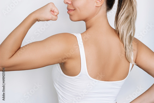 Young strong fit blonde woman standing with her back raising arm and showing bicep isolated on a white background. Feminism, girl power, equal women's rights, independence, sports concept
