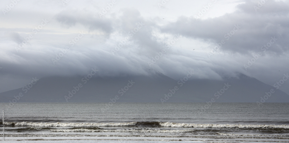 A mountain is obscured by morning storm clouds, which produce a blueish-gray scene with soaring birds, a dramatic sky, and crashing waves. Inch Beach, Dingle Peninsula, County Kerry, Ireland A mountain is obscured by morning storm clouds, which produce a blueish-gray scene with soaring birds, a dramatic sky, and crashing waves. Inch Beach, Dingle Peninsula, County Kerry, Ireland