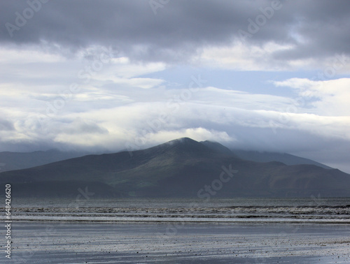 A small gap in the heavy clouded sky highlights the lush and green slope of a mountain range emerging from dramatic North Atlantic Ocean water. Inch Beach, Dingle Peninsula, County Kerry, Ireland