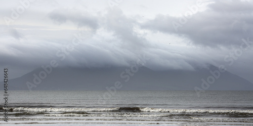 A mountain is obscured by morning storm clouds, which produce a blueish-gray scene with soaring birds, a dramatic sky, and crashing waves. Inch Beach, Dingle Peninsula, County Kerry, Ireland