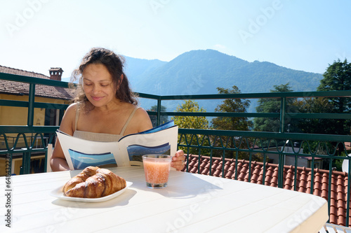 Happy woman reading magazine while taking her breakfast in the summer terrace, against the beautiful nature background