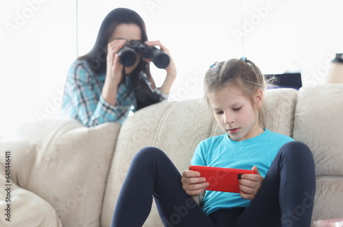 Mom controls her daughter with phone through binoculars. Best parental controls and smartphone apps concept photo
