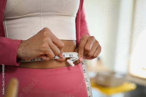 Midsection of slim woman in sports clothes measuring waist with tape at home. Sports dieting, weight loss and healthy lifestyle concept