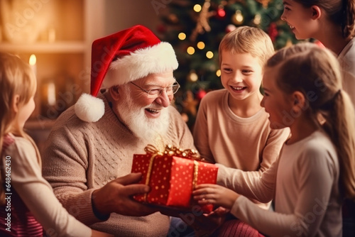 Happy satisfied grandfather wearing red santa hat giving his cute smiling little grandchildren christmas gifts