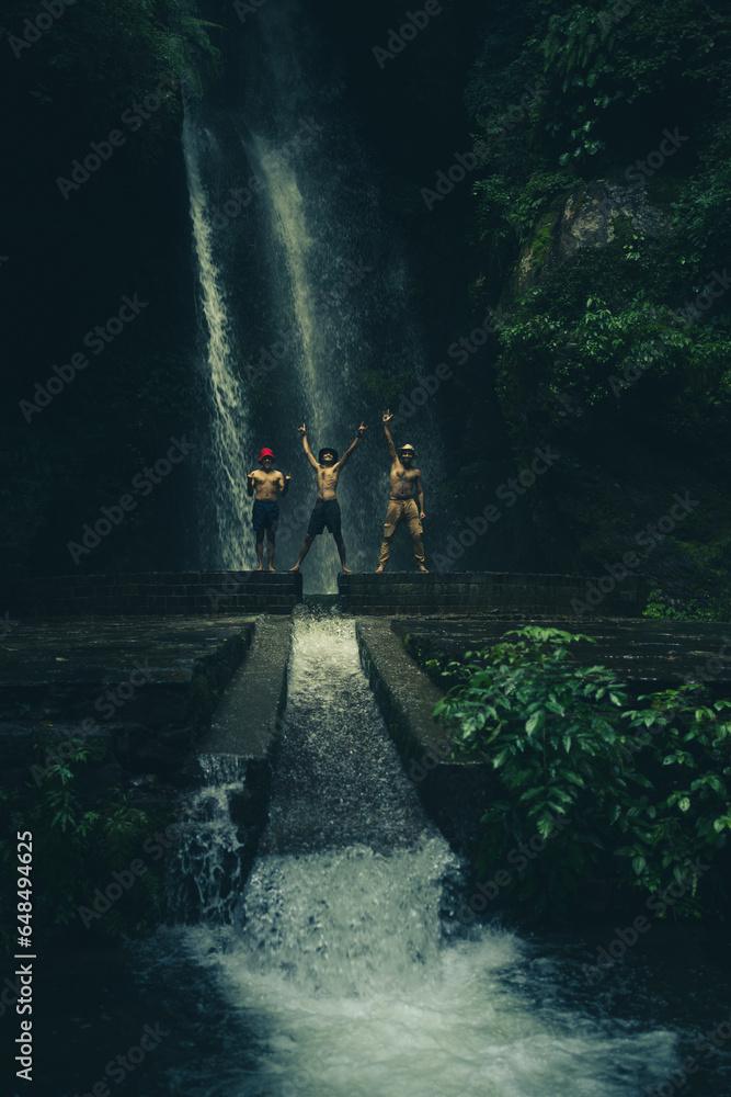 Obraz premium A man standing on a rock in front of a tall waterfall in the mountains, set in the tropical landscape of Jibhi Waterfall, Himachal Pradesh, India.