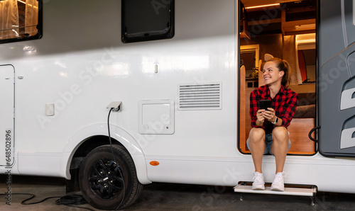 Happy young woman sitting in camper van uses her cell phone while van is charging on camper park. House on wheels. RV rental.
